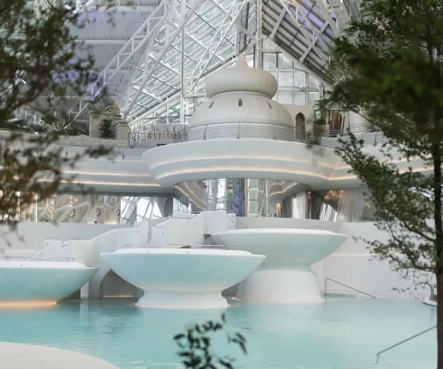 Una zona de piscina coberta amb aigua blava clara, envoltada d'arquitectura moderna blanca amb grans estructures en forma de bol i un edifici amb cúpula. Un complex sostre de vidre i bigues és visible a dalt, amb una mica de fullatge verd en primer pla.