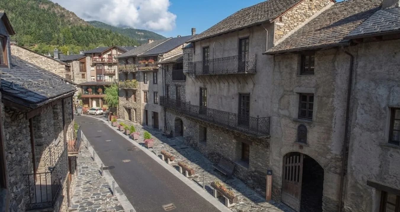 Casa Museu d'Areny-Plandolit. Una calle estrecha y pavimentada serpentea a través de un pueblo de montaña histórico, flanqueada por edificios tradicionales de piedra con techos oscuros y balcones de hierro forjado. Montañas verdes y boscosas se elevan al fondo bajo un cielo parcialmente nublado.