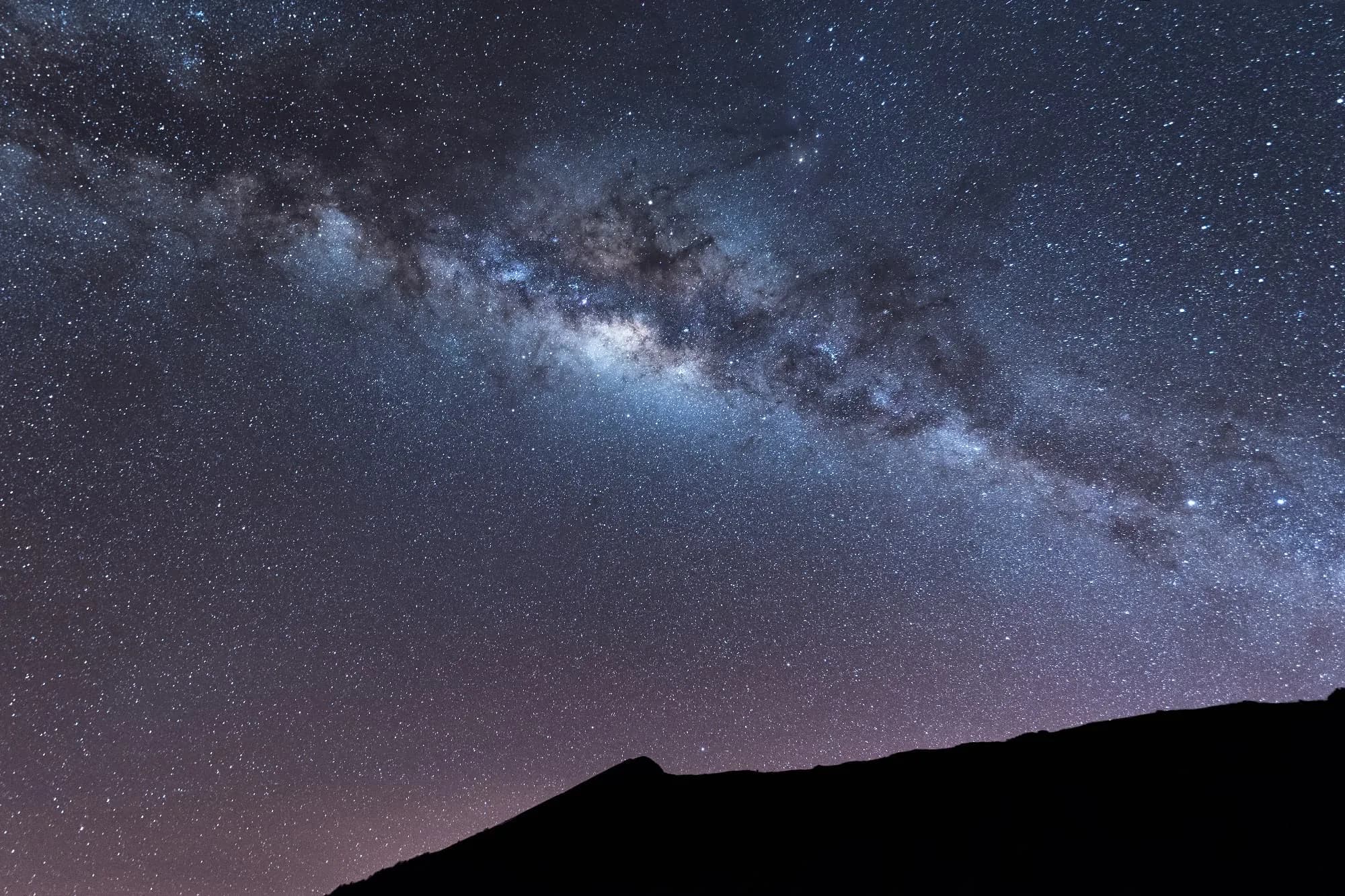La Via Làctia s'arqueja a través d'un cel nocturn ple d'estrelles, amb la silueta fosca d'una muntanya en primer pla.
