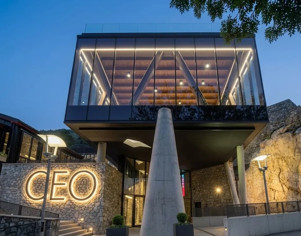 Un edifici modern amb un pis superior de vidre il·luminat al capvespre, amb un gran rètol lluminós de "CEO" en una paret de pedra a sota.