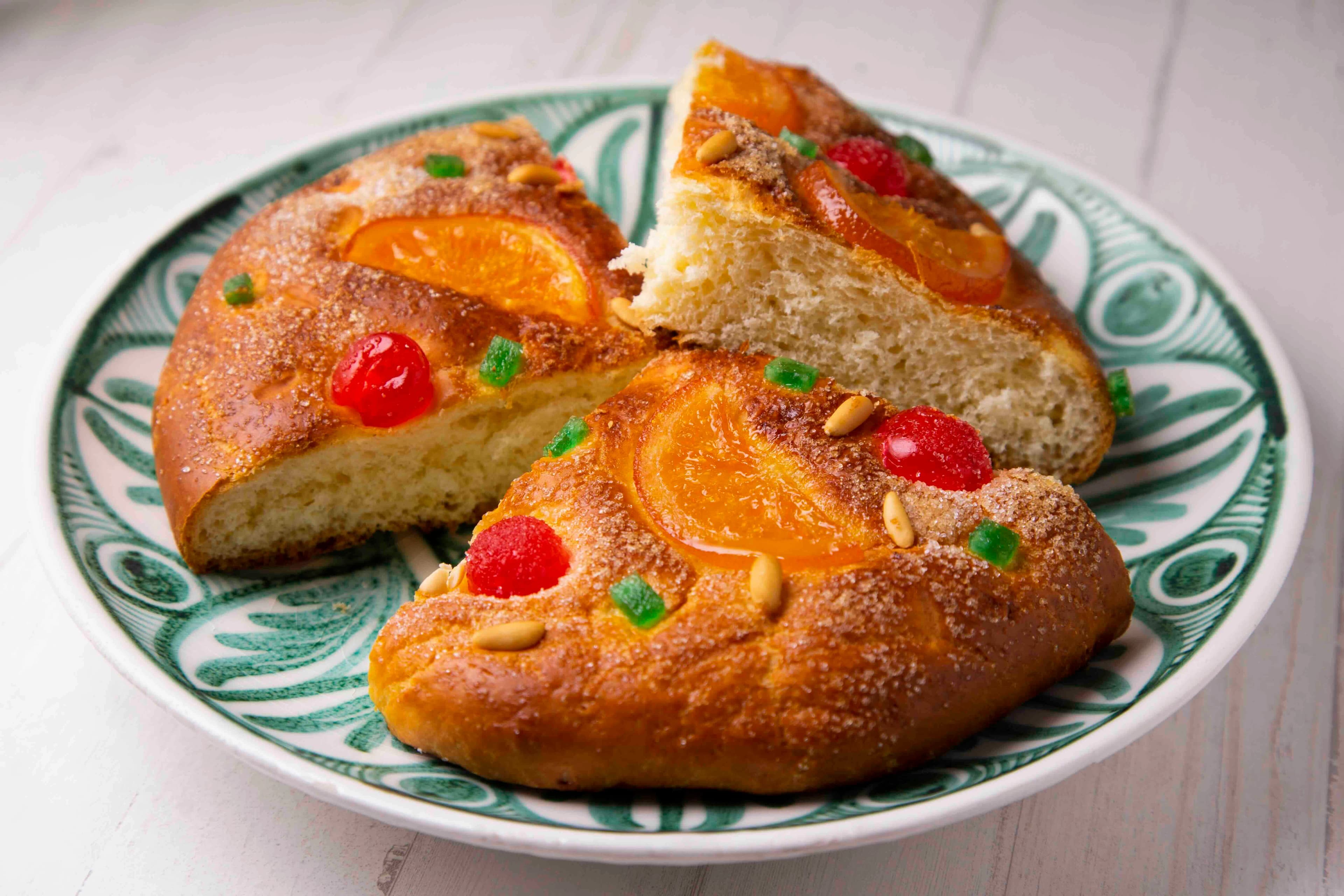 Tres trossos de Coca de Sant Joan, un pa dolç tradicional decorat amb taronja confitada, cireres vermelles, fruita verda i pinyons, sobre un plat blanc i verd.