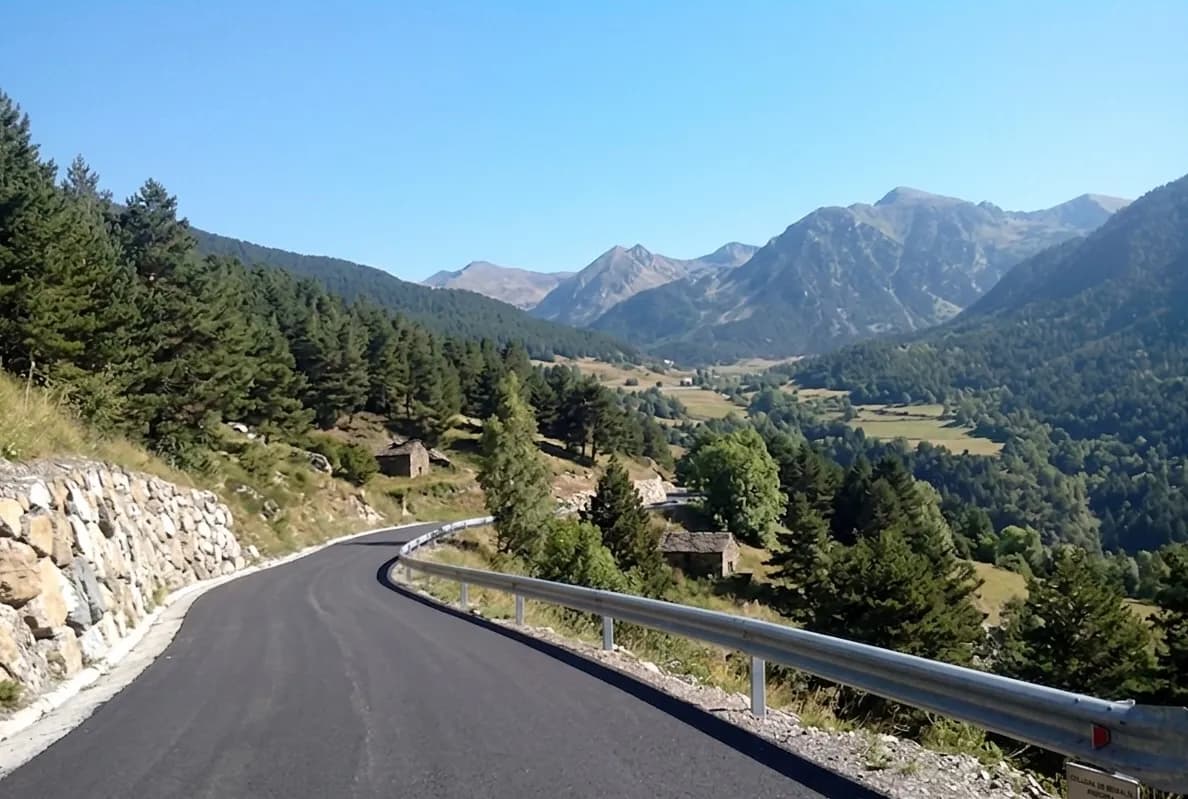 A winding asphalt road with a stone wall on the left and a guardrail on the right, leading through a mountainous landscape with pine forests, green valleys, and distant peaks under a clear blue sky. Small stone buildings are nestled among the trees.