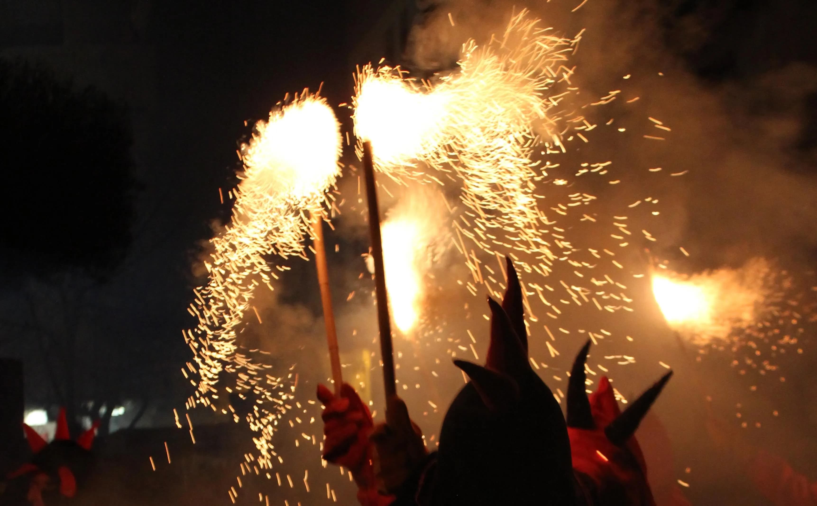 Siluetes de persones amb disfresses de diable sostenen bengales que emeten espurnes daurades brillants i fum contra un cel nocturn fosc durant una celebració de Correfoc.