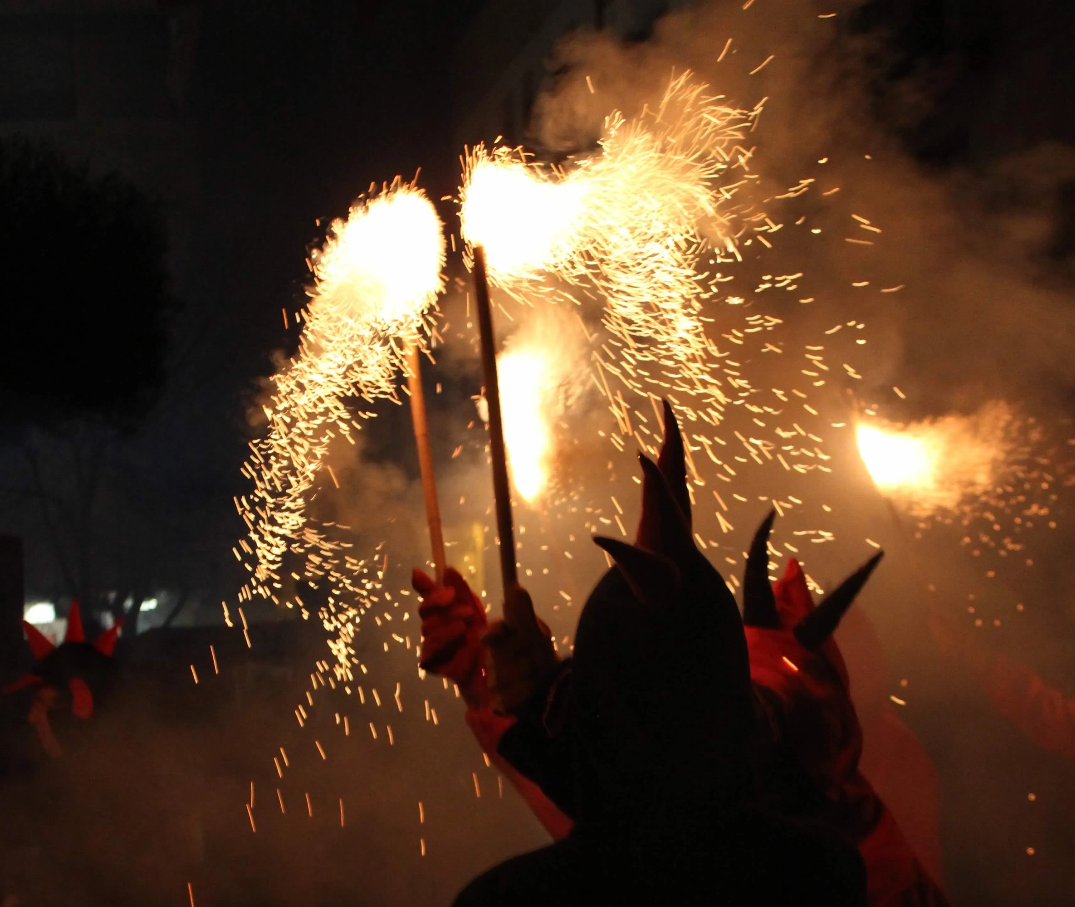 Una persona amb una disfressa fosca i amb banyes sosté dues bengales que emeten espurnes daurades brillants i fum a la nit. Les espurnes il·luminen l'entorn fosc i el fum, creant una escena dinàmica.