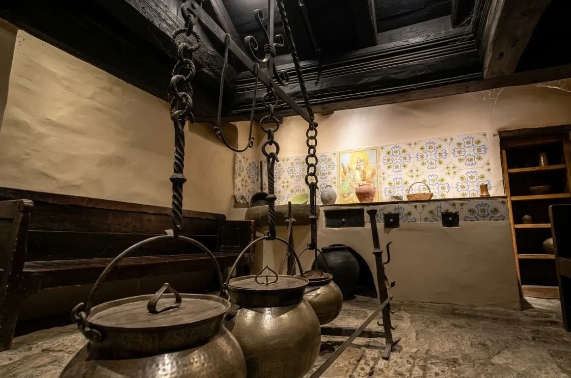 Una cuina rústica i tradicional amb un gran suport de ferro forjat que sosté diverses olles de coure en primer pla. El fons presenta un taulell de rajoles amb art religiós, un banc de fusta i bigues de sostre exposades.