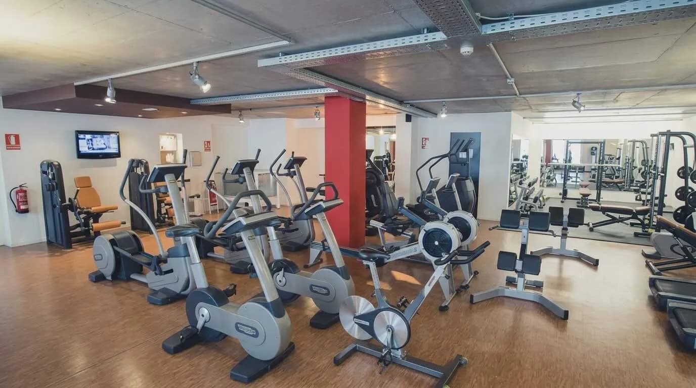 Un gimnàs ben equipat amb una varietat de màquines d'exercici, incloent bicicletes estàtiques, el·líptiques i màquines de rem en primer pla. Al fons es veuen equips d'aixecament de peses i miralls, tot sobre un terra de fusta.