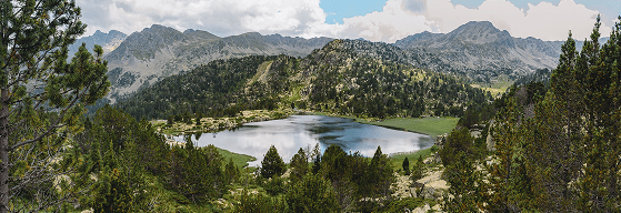 Vista panorámica de un sereno lago de montaña rodeado de bosques de pinos y picos escarpados bajo un cielo parcialmente nublado. El agua tranquila refleja el paisaje natural.