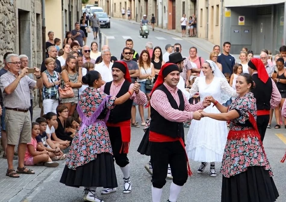 Un grup de persones amb vestits folklòrics tradicionals balla en un carrer, agafats de la mà. Els espectadors s'alineen als costats del carrer, observant l'actuació.