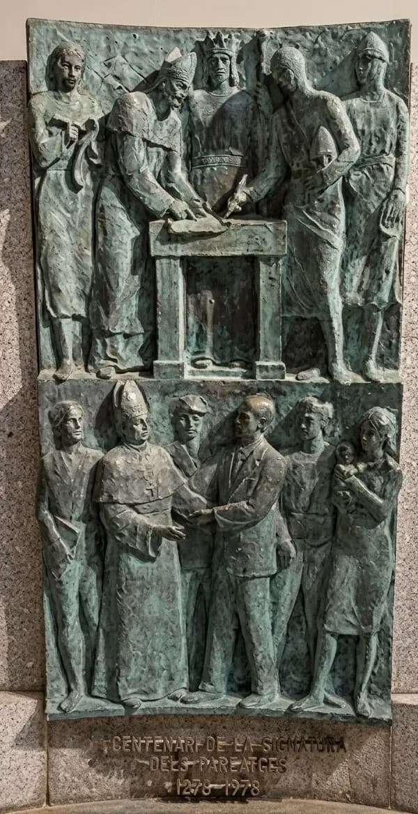 Una escultura en relleu de bronze que commemora els Pareatges, mostrant una escena superior de figures històriques signant un document i una escena inferior d'un bisbe donant la mà a un home de vestit, envoltat de gent. La inscripció diu "CENTENARI DE LA SIGNATURA DELS PAREATGES 1278-1978".
