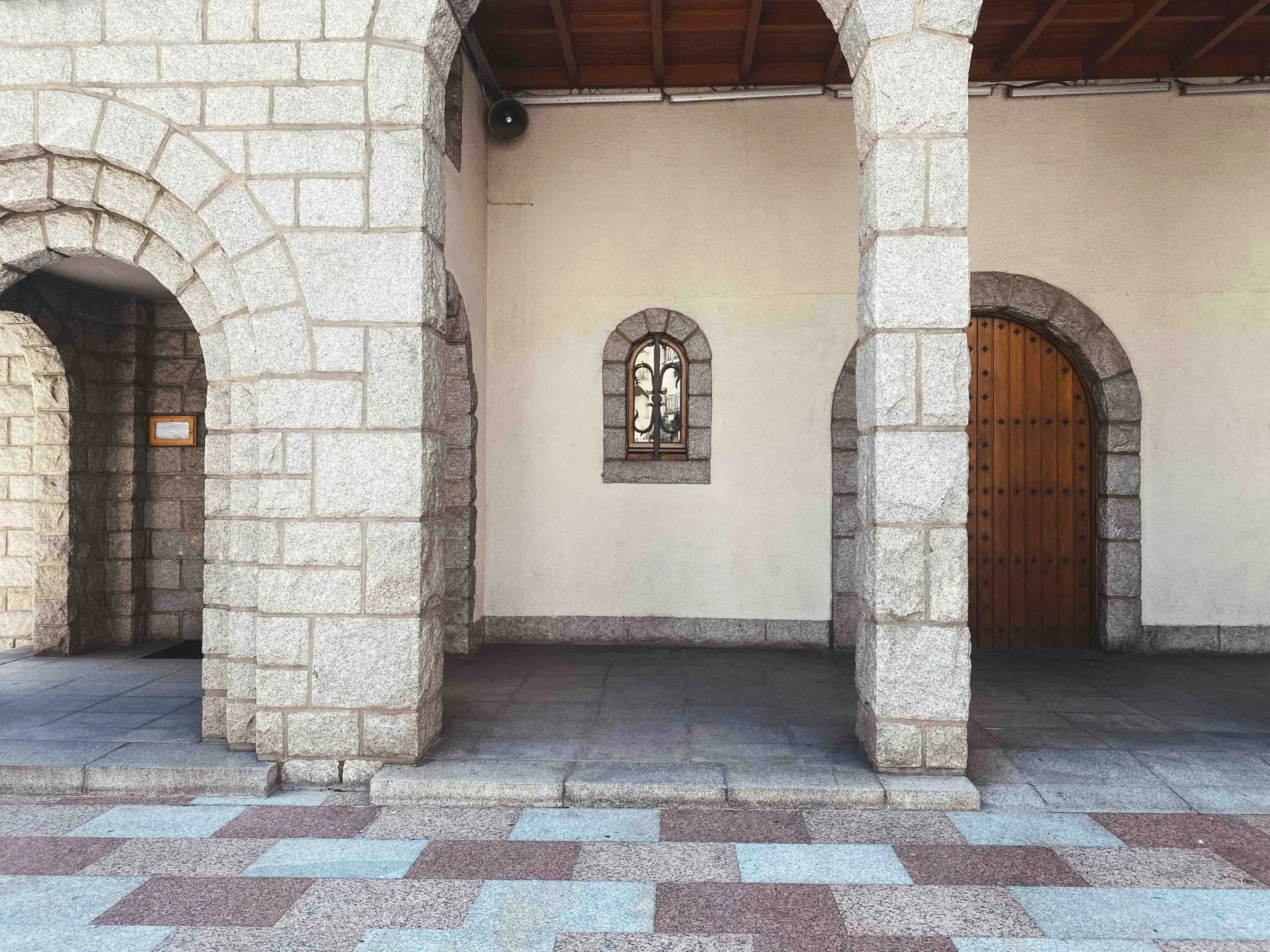 Una façana d'edifici amb arcs i columnes de pedra de color clar, que presenta una petita finestra arquejada amb una reixa de metall i una porta de fusta arquejada. El terra està pavimentat amb rajoles estampades en gris, marró i blau clar.