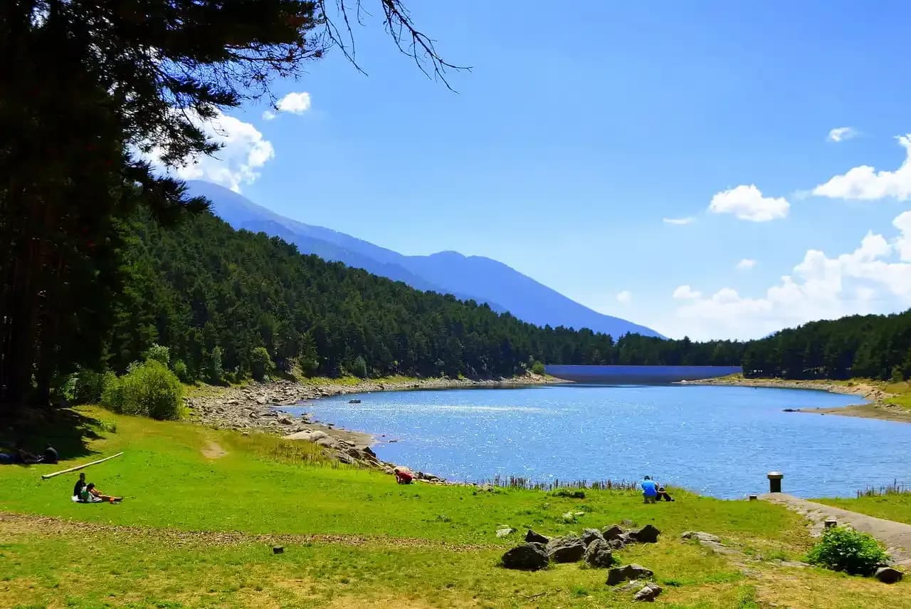 Una vista panoràmica d'un llac blau tranquil envoltat per un dens bosc verd i muntanyes llunyanes sota un cel blau brillant. Algunes persones es relaxen a la riba herba en primer pla.