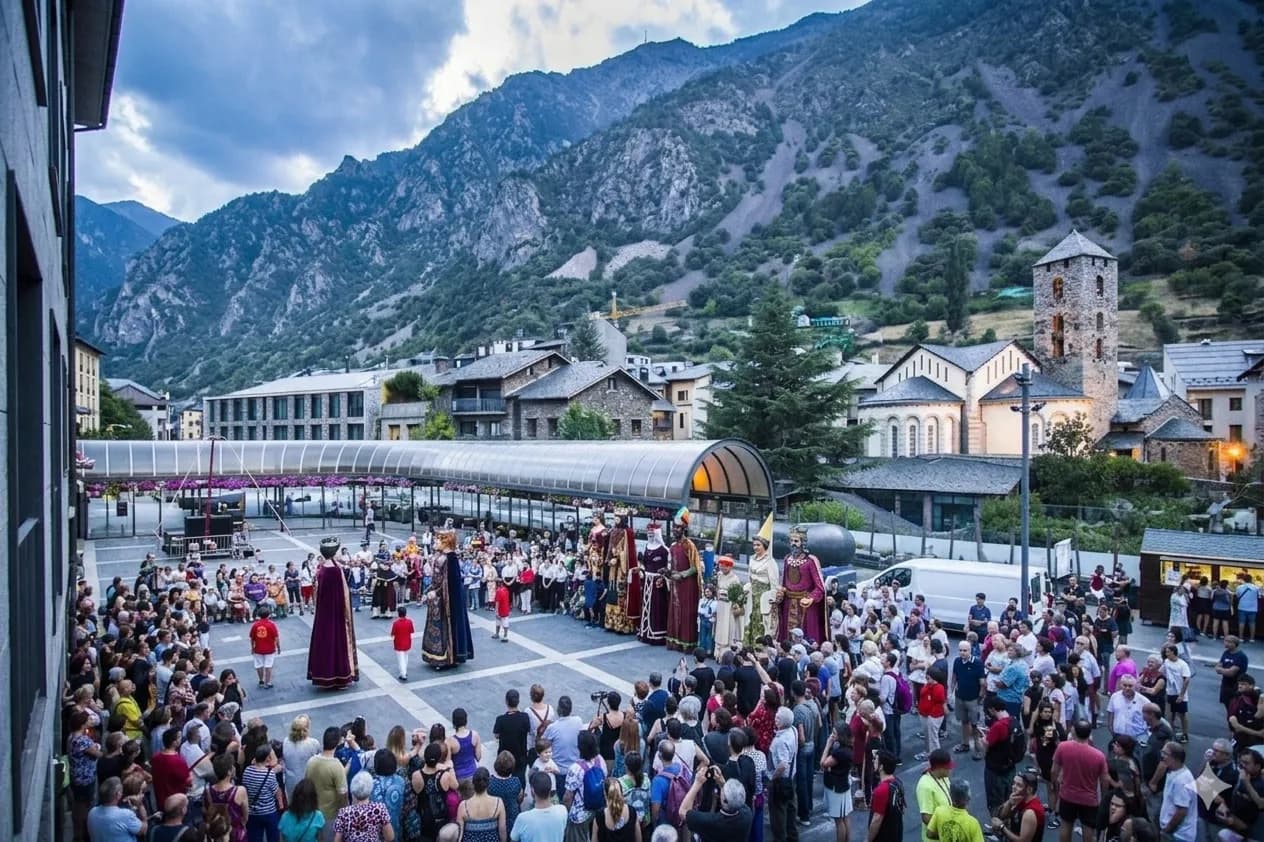Una gran multitud es reuneix en una plaça del poble per veure una desfilada de figures gegants amb vestits tradicionals. Muntanyes i edificis tradicionals, incloent una església amb una torre de pedra, emmarquen l'escena sota un cel ennuvolat.
