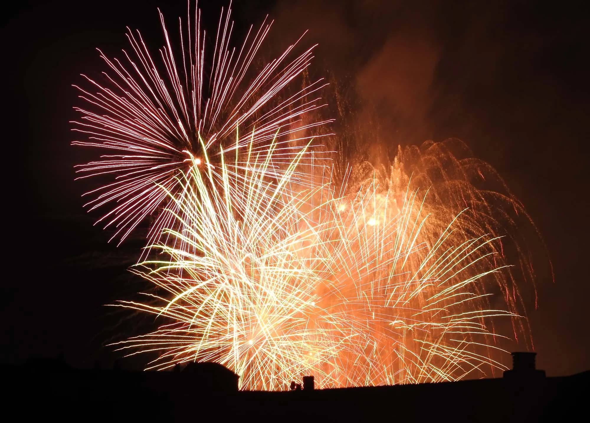 Un espectacular espectacle de focs artificials il·lumina el cel nocturn amb explosions de llum vermella, groga i taronja sobre una silueta fosca d'edificis.
