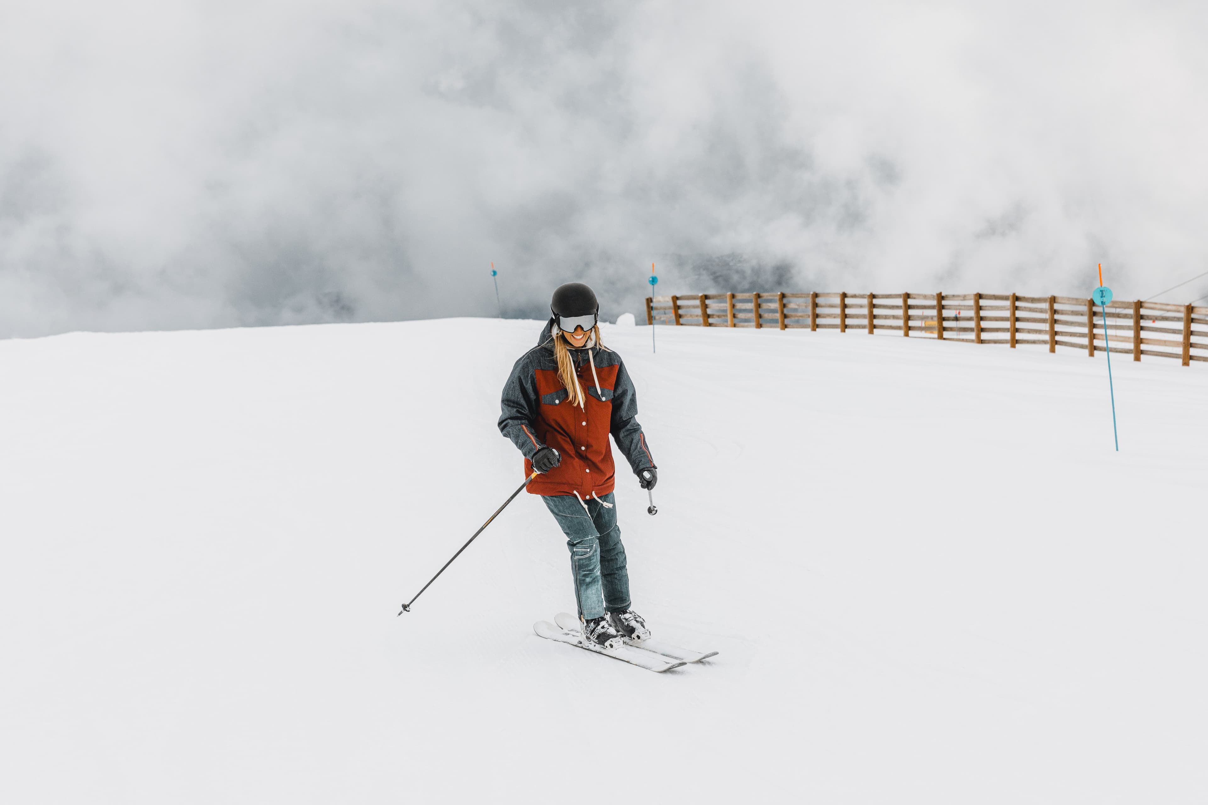 Mujer sonriente esquiando en el pirineo en una pista nevada en un día nublado, vistiendo una chaqueta roja y gris con casco negro.