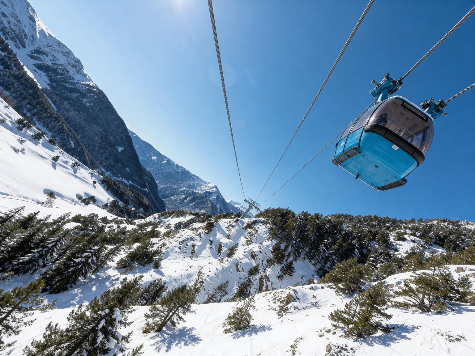 Una cabina blava de telecabina llisca sobre un vessant de muntanya nevat amb pins sota un cel blau clar. El sol brilla a la part superior esquerra.
