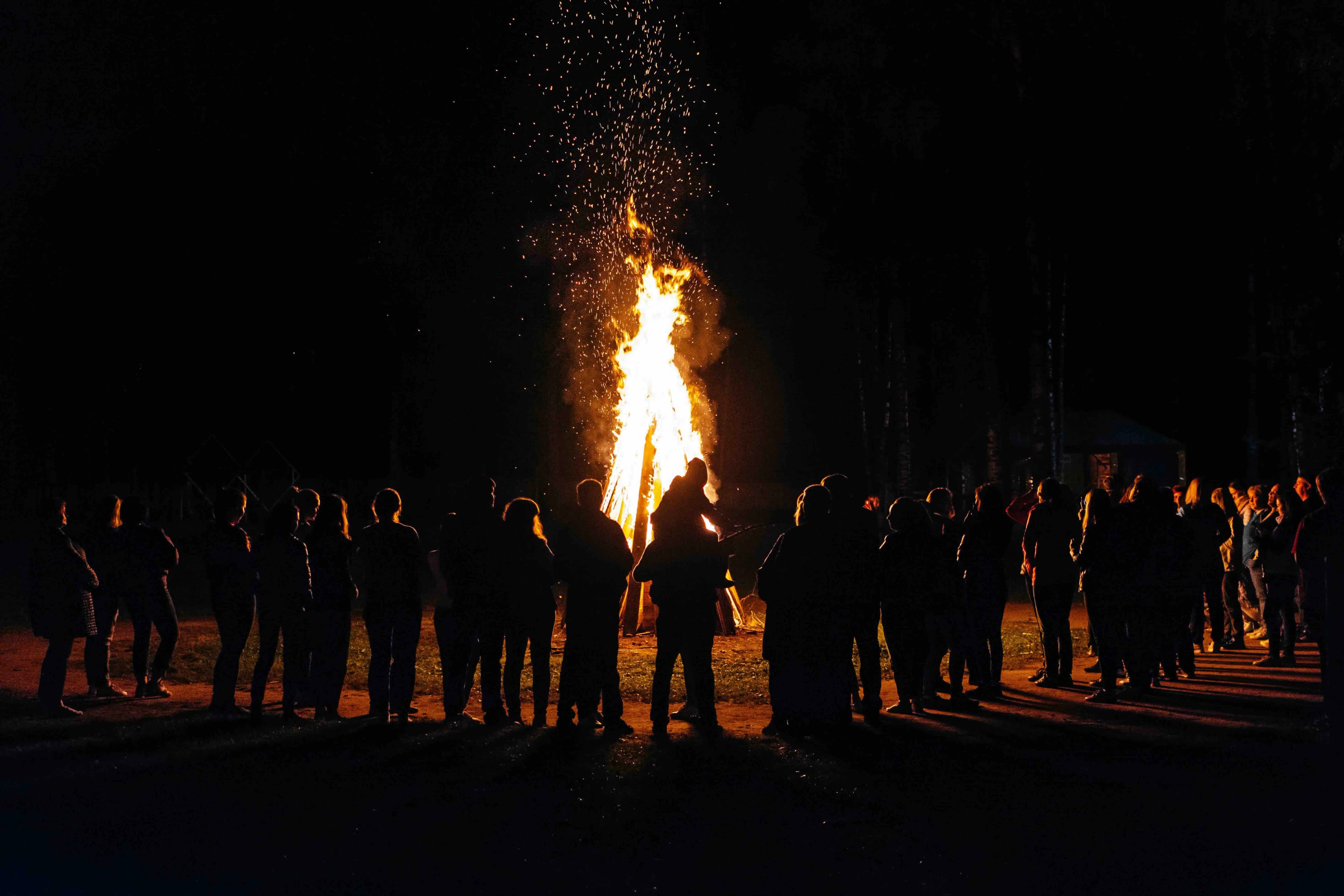 Un grup de persones en silueta es reuneix al voltant d'una gran foguera brillant a la nit. Espurnes volen cap amunt des del foc, i una persona porta un nen a les espatlles.