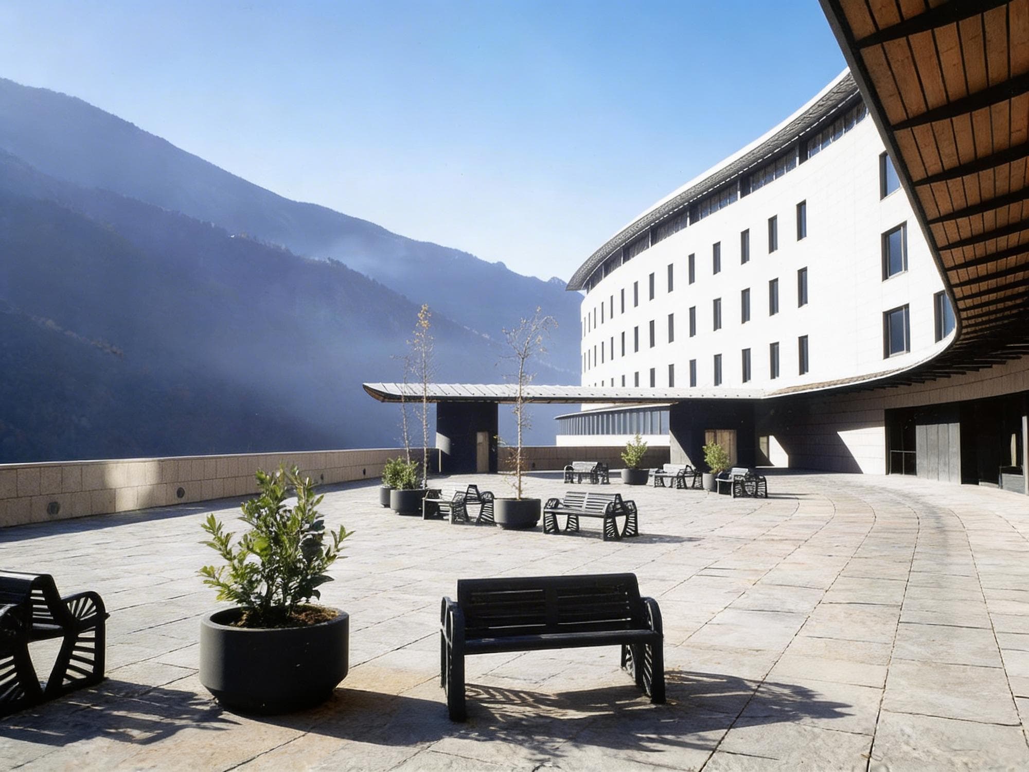 Un edifici modern i corbat de façana blanca amb moltes finestres domina un ampli pati pavimentat amb bancs negres i plantes en testos. Muntanyes boiroses s'eleven al fons sota un cel blau clar.