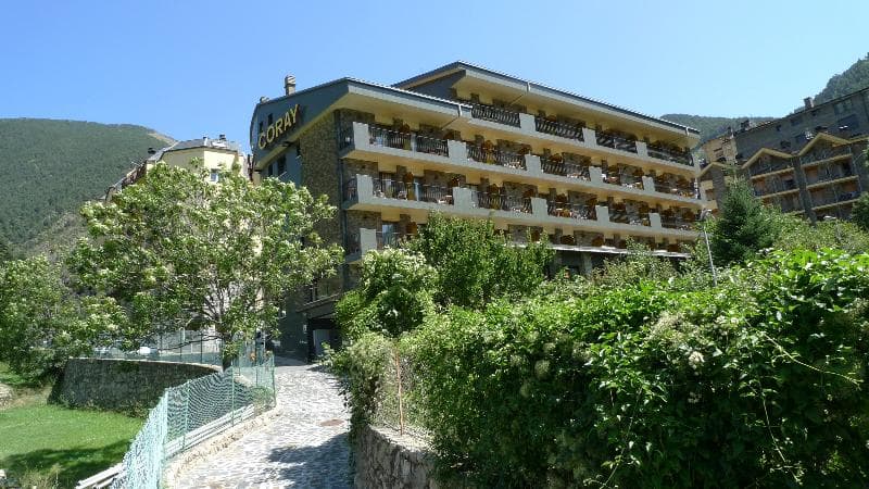 Un edifici d'hotel de diversos pisos anomenat "CORAY" amb balcons es troba en un vessant, envoltat d'exuberants arbres i arbustos verds sota un cel blau clar. Un camí empedrat condueix cap a l'edifici.