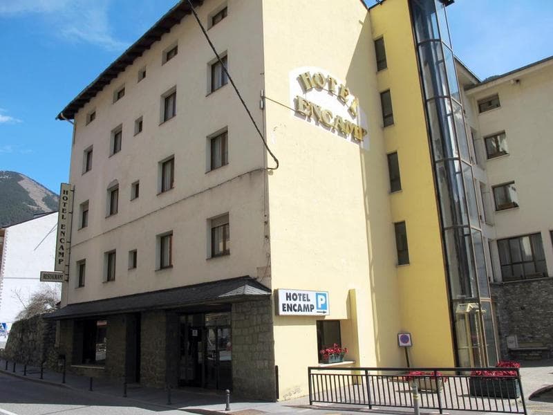 Un edifici d'hotel de diverses plantes anomenat "Hotel Encamp" amb una façana de color clar, un ascensor de vidre prominent i rètols de restaurant i aparcament. Es veuen muntanyes al fons.