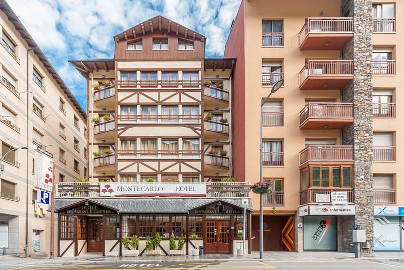 Vista a peu de carrer de l'Hotel Montecarlo, un edifici de diverses plantes amb una façana distintiva marró i crema amb bigues de fusta i balcons. Està flanquejat per altres edificis d'apartaments, amb un carrer i diversos rètols visibles en primer pla.