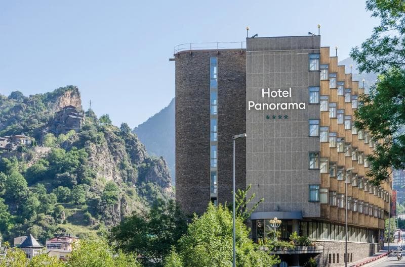 Un gran edificio de hotel, "Hotel Panorama" con cuatro estrellas, se alza prominentemente contra un telón de fondo de exuberantes montañas verdes y un cielo despejado. Árboles y otros edificios son visibles en las laderas.