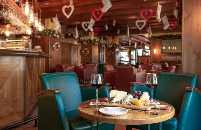 Un acogedor bar o salón con una mesa de madera en primer plano que presenta una tabla de quesos, uvas y dos copas de vino tinto. La sala está decorada con corazones rojos, calcetines blancos y guirnaldas, creando un ambiente festivo.