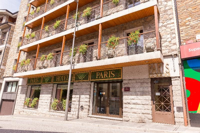 L'exterior de l'Hotel Paris, un edifici de diverses plantes amb façana de pedra i balcons adornats amb plantes i flors. L'entrada de l'hotel disposa de portes de vidre i el nom 'PARIS HOTEL' es mostra de manera destacada.