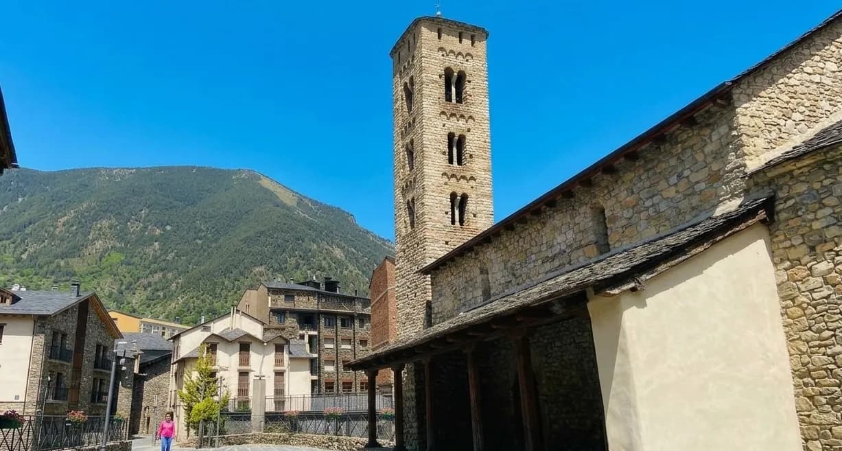 Una alta torre campanar rectangular de pedra s'alça al costat d'un edifici de pedra amb un passadís cobert, sota un cel blau clar. Al fons, muntanyes verdes s'eleven sobre cases de pedra tradicionals.
