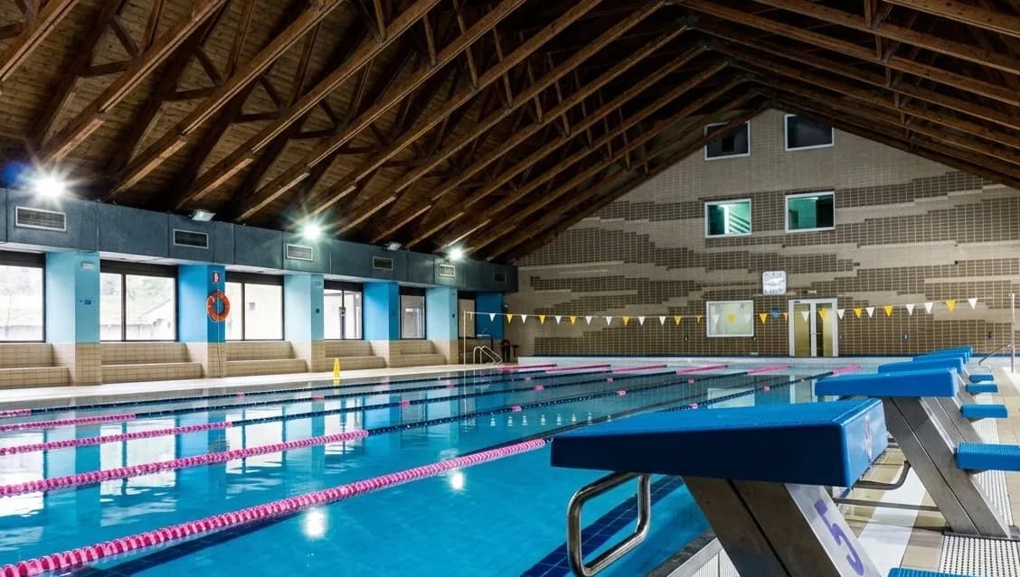 Una piscina coberta amb aigua blava, separadors de carril roses i blocs de sortida blaus. La zona de la piscina presenta grans finestrals, parets blau clar i un sostre alt amb bigues de fusta.
