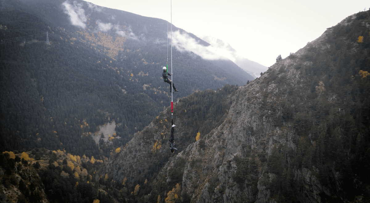Dues persones estan suspeses per cordes a gran alçada sobre una profunda vall muntanyosa i boscosa sota un cel ennuvolat. Una persona està dreta, mentre que una altra penja cap per avall a sota, participant en un esport extrem com el puenting o el ràpel.