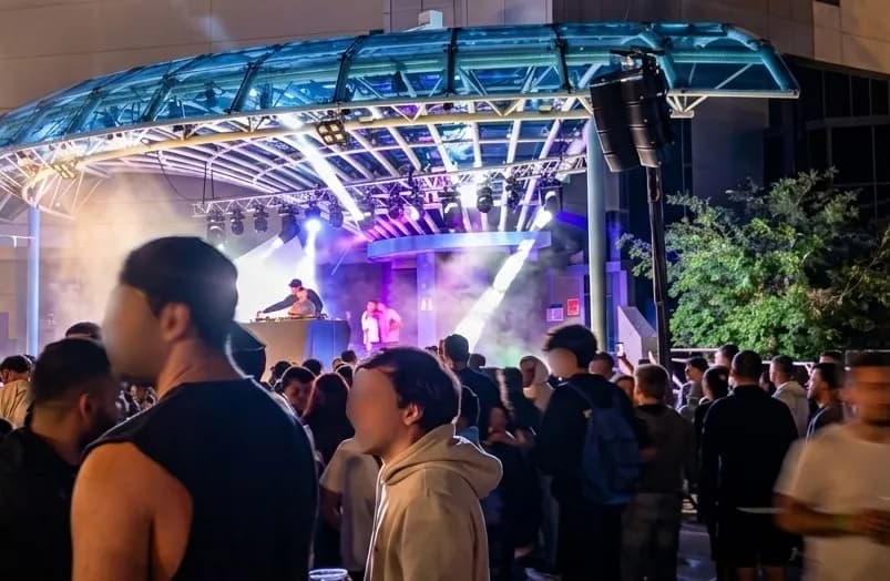 Un concert nocturn a l'aire lliure amb un DJ i un artista a l'escenari, envoltat per una multitud de persones i llums brillants.