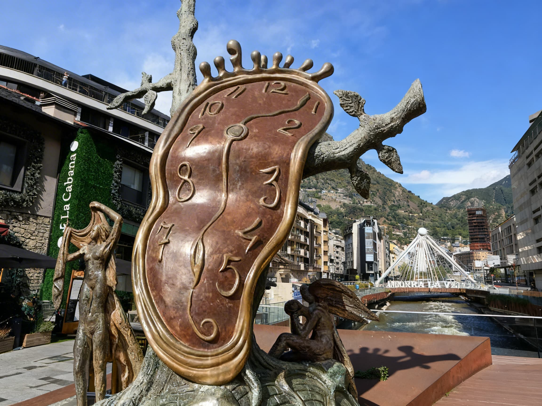 Una gran escultura de bronce de Dalí, "Noblesse du Temps", con un reloj derretido y otras figuras, se encuentra en Andorra la Vella. Al fondo se ven edificios urbanos, un río con un puente moderno y montañas bajo un cielo azul.