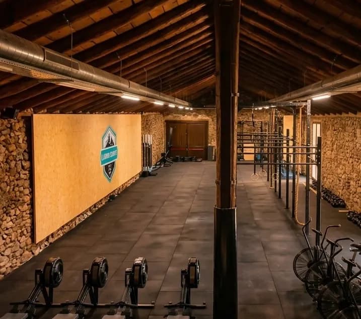 Vista interior d'un gimnàs de CrossFit amb sostre alt de bigues de fusta, parets de pedra i terra de goma fosc. L'equip inclou màquines de rem, una gran estructura metàl·lica amb anelles, bicicletes de aire i peses, amb un logotip de 'LLARD CROSSFIT' en un panell de fusta.
