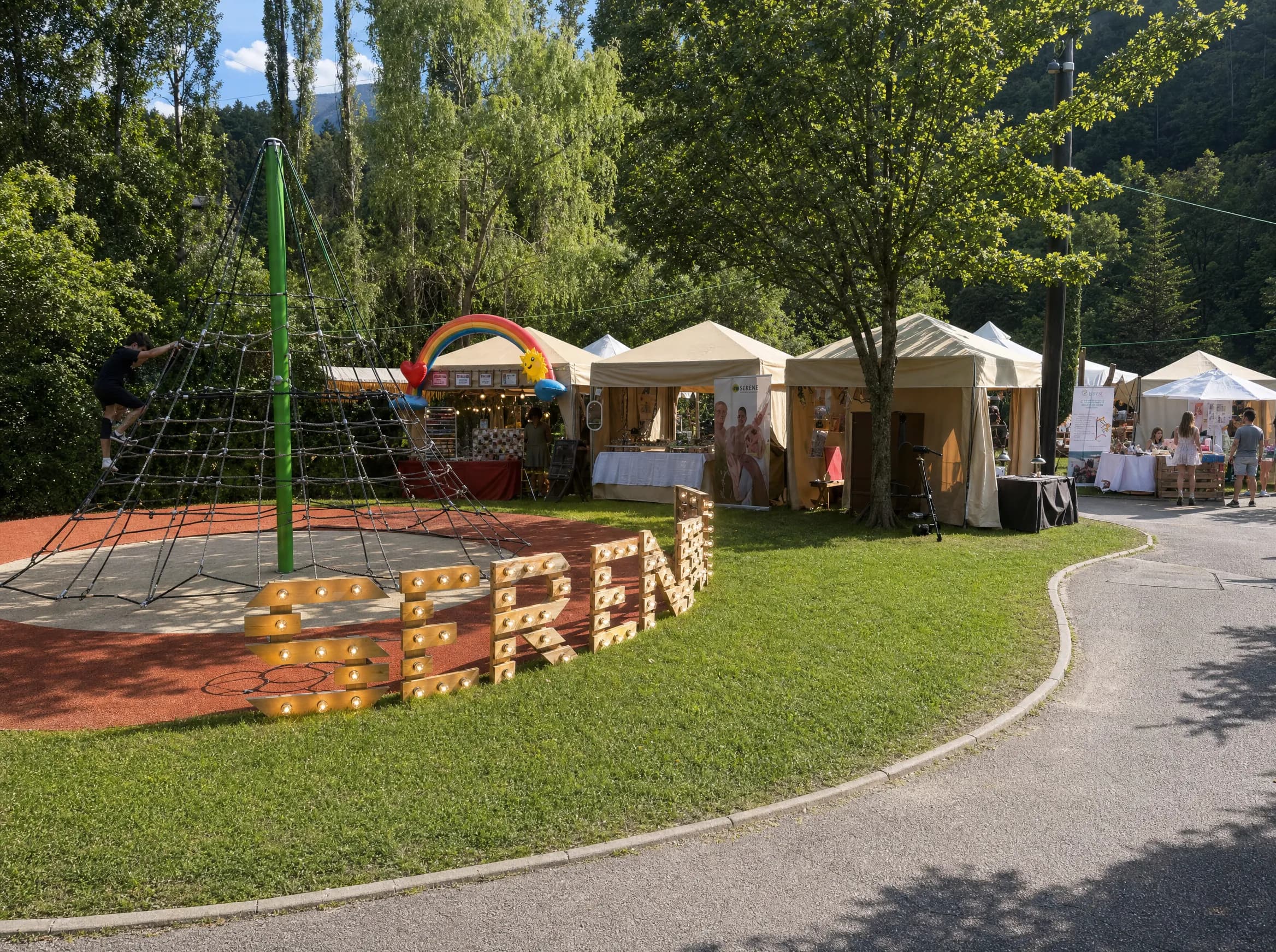 Una escena vibrant a l'aire lliure amb un parc infantil que inclou una estructura d'escalada de corda i una persona escalant-la. Al fons, diverses parades de mercat estan muntades, una decorada amb un arc de Sant Martí, i la gent està mirant.