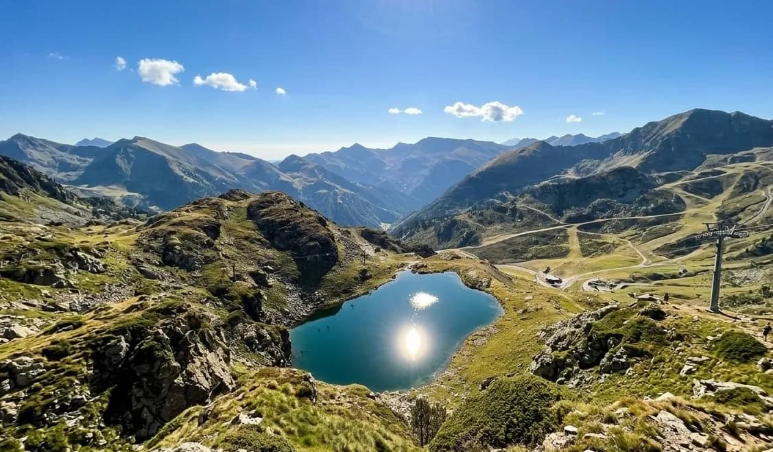 Una vista aèria d'un llac de muntanya amb el sol reflectint-se brillantment a la seva superfície, envoltat de vessants rocosos i herbosos i muntanyes distants sota un cel blau clar. Una torre de telecadira és visible a la dreta.