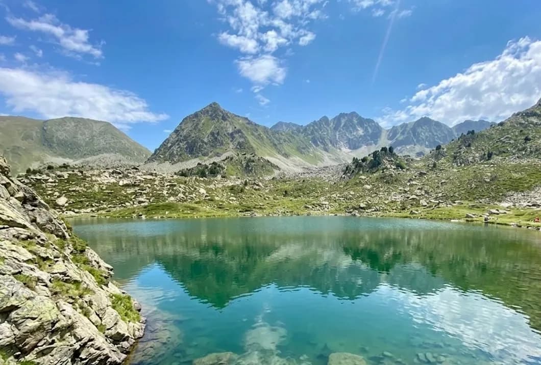 Un llac de muntanya de color turquesa clar reflecteix cims rocosos i verds sota un cel blau amb núvols blancs, amb vegetació verda i roques a la seva vora.