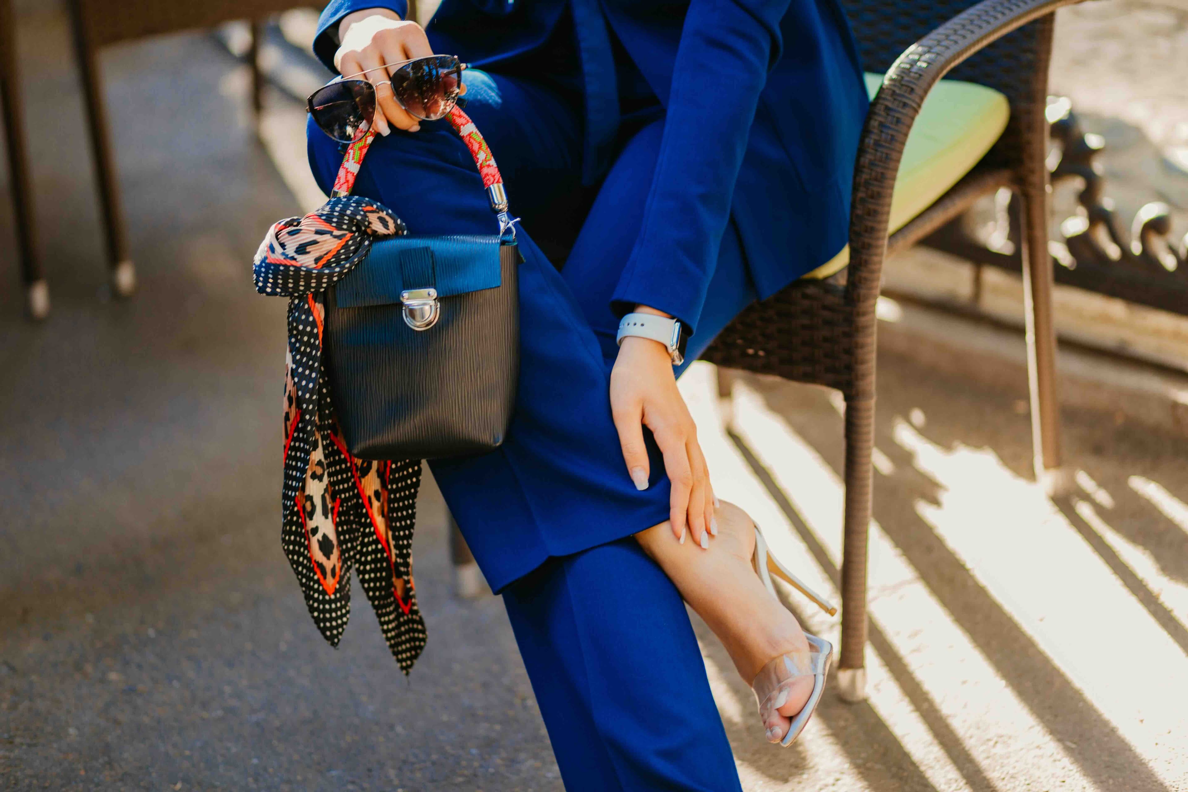 Una persona con un traje azul sostiene un bolso negro adornado con un pañuelo colorido y gafas de sol, mientras está sentada en una silla de mimbre. Se ve un reloj en su muñeca y lleva tacones altos.