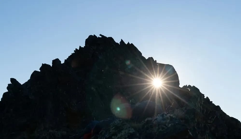 El sol crea un efecte d'estrella brillant mentre treu el cap per darrere d'un pic de muntanya fosc i dentat, retallat contra un cel blau clar.
