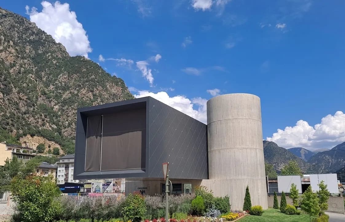 Un edifici modern amb una secció rectangular gris fosc i una secció cilíndrica gris clar, situat davant d'una gran muntanya verda sota un cel blau amb núvols blancs. En primer pla hi ha jardineria amb arbustos i flors.