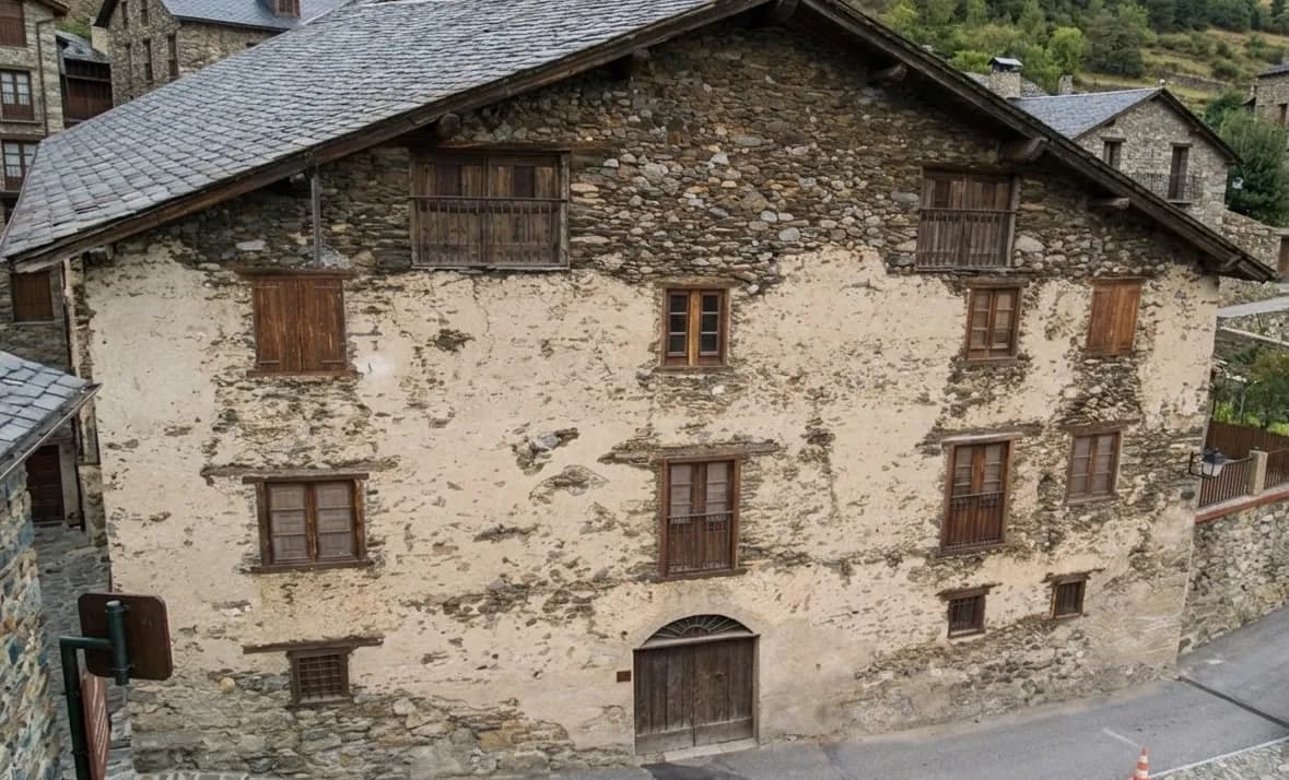 Un antiguo edificio de varias plantas de piedra y estuco con tejado de pizarra gris y numerosas ventanas de madera, algunas con contraventanas o pequeños balcones, y una gran puerta de madera arqueada en la base, situado en un pueblo rústico.
