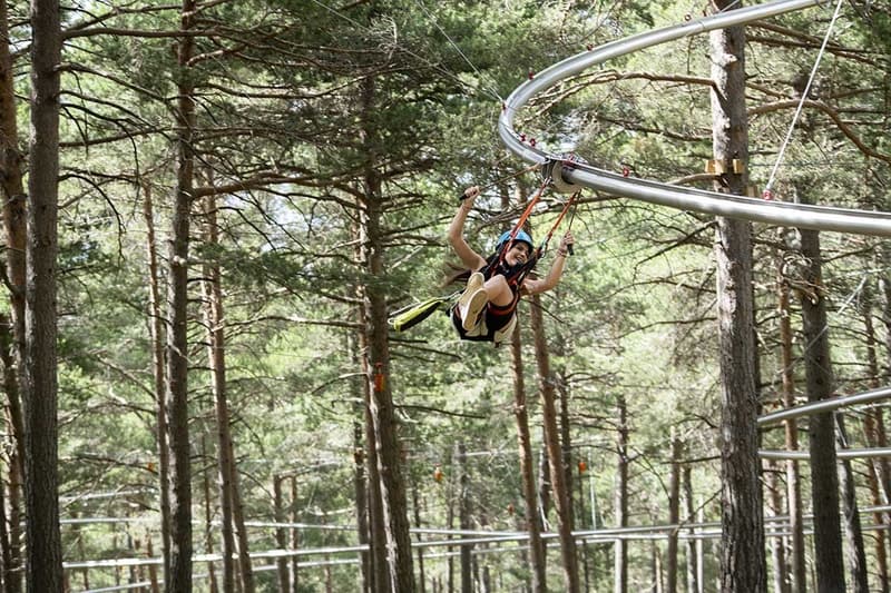 Una persona con casco y arnés sonríe mientras está suspendida de una atracción aérea, deslizándose por un denso bosque de pinos en una pista metálica curva.