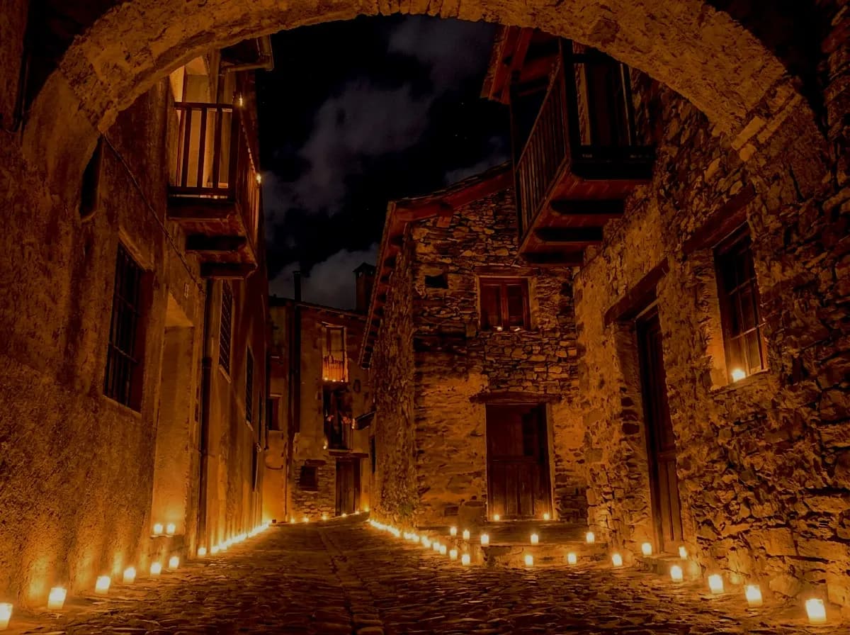 Un carrer estret i antic de pedra de nit, il·luminat per nombroses espelmes petites que voregen el camí i es col·loquen als ampits de les finestres i esglaons. Edificis de pedra amb balcons de fusta flanquegen el carrer, que està arquejat a la part superior.