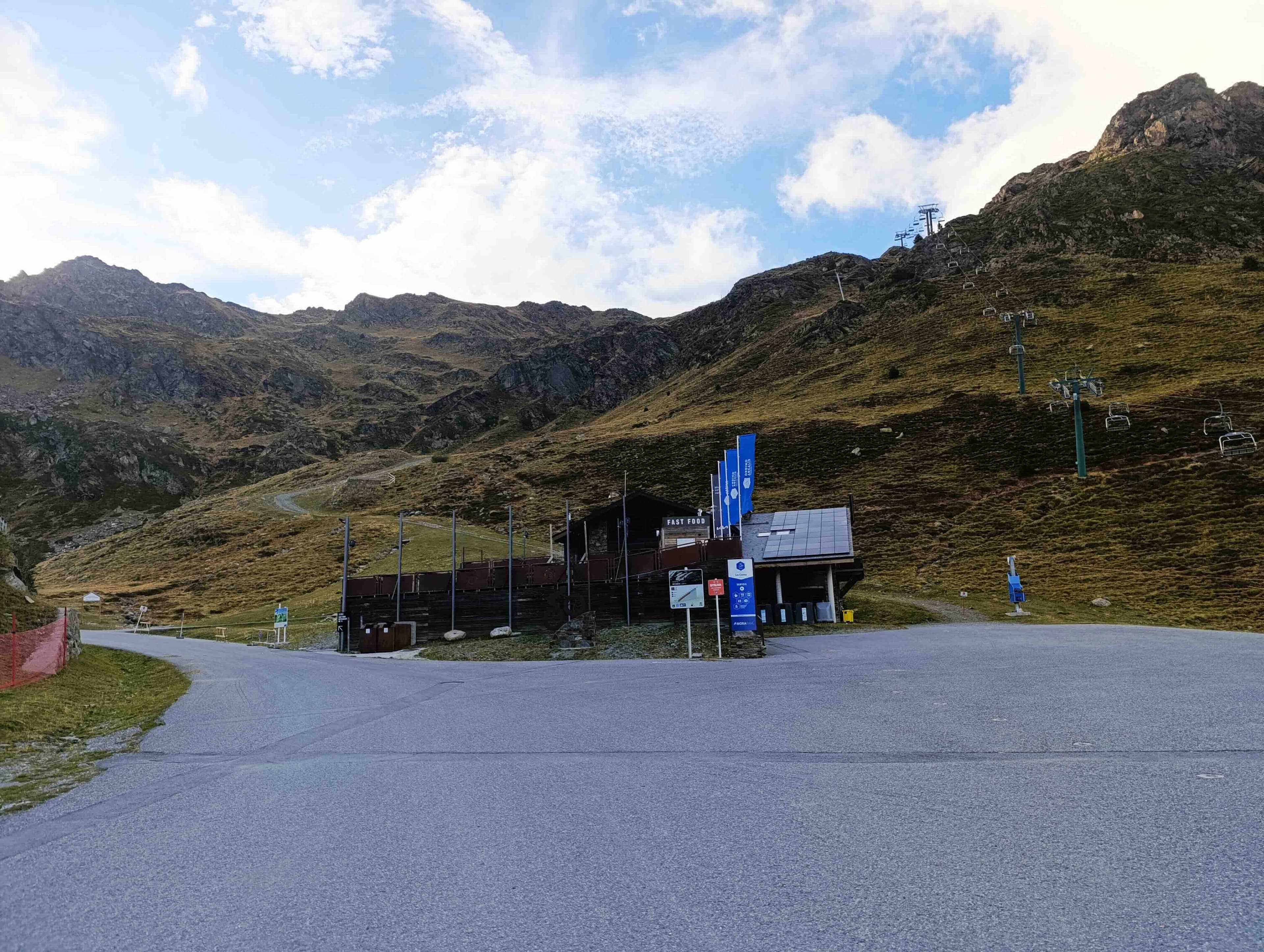 Un edifici d'estació d'esquí amb panells solars i banderes es troba al costat d'una carretera asfaltada al peu d'una muntanya rocosa, on els telecadires ascendeixen sota un cel parcialment ennuvolat.