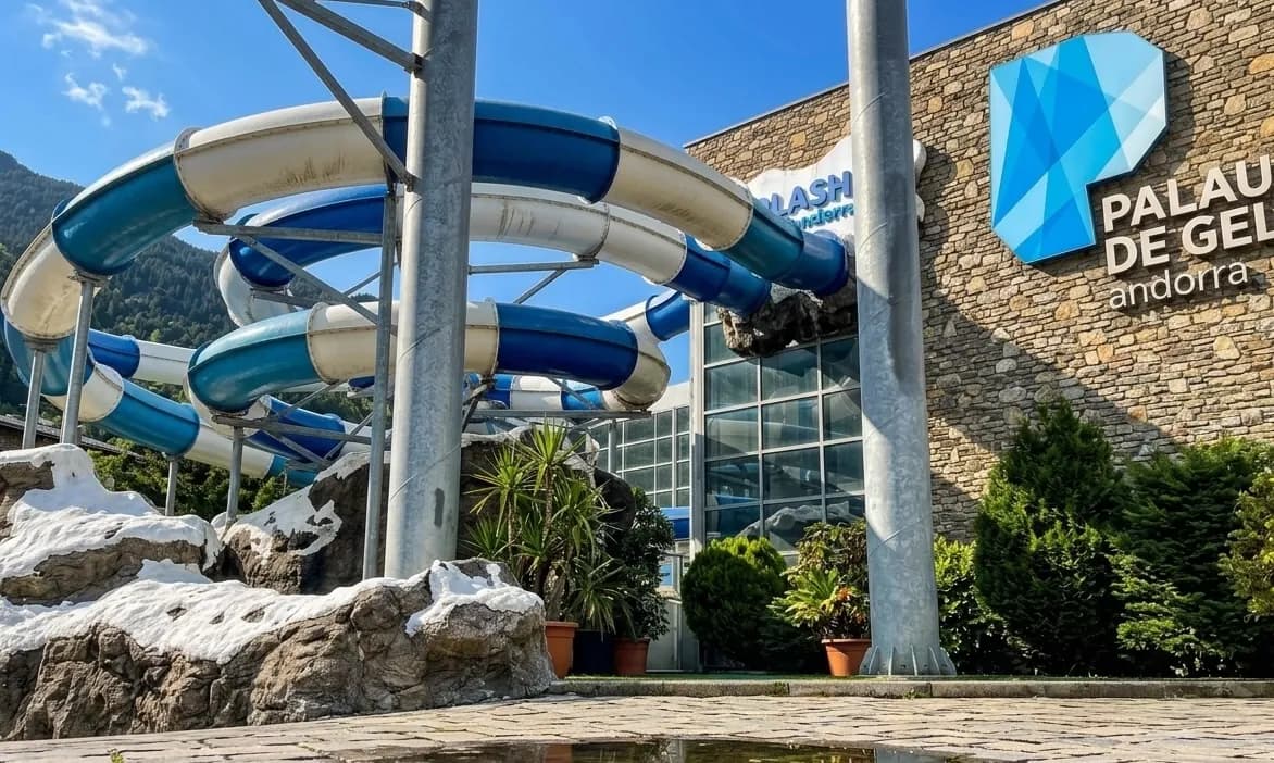 Un tobogan d'aigua blau i blanc serpenteja al costat d'un edifici de pedra amb els rètols 'Palau de Gel Andorra' i 'Splash Andorra'. A la base hi ha plantes verdes i formacions rocoses artificials, amb muntanyes al fons.