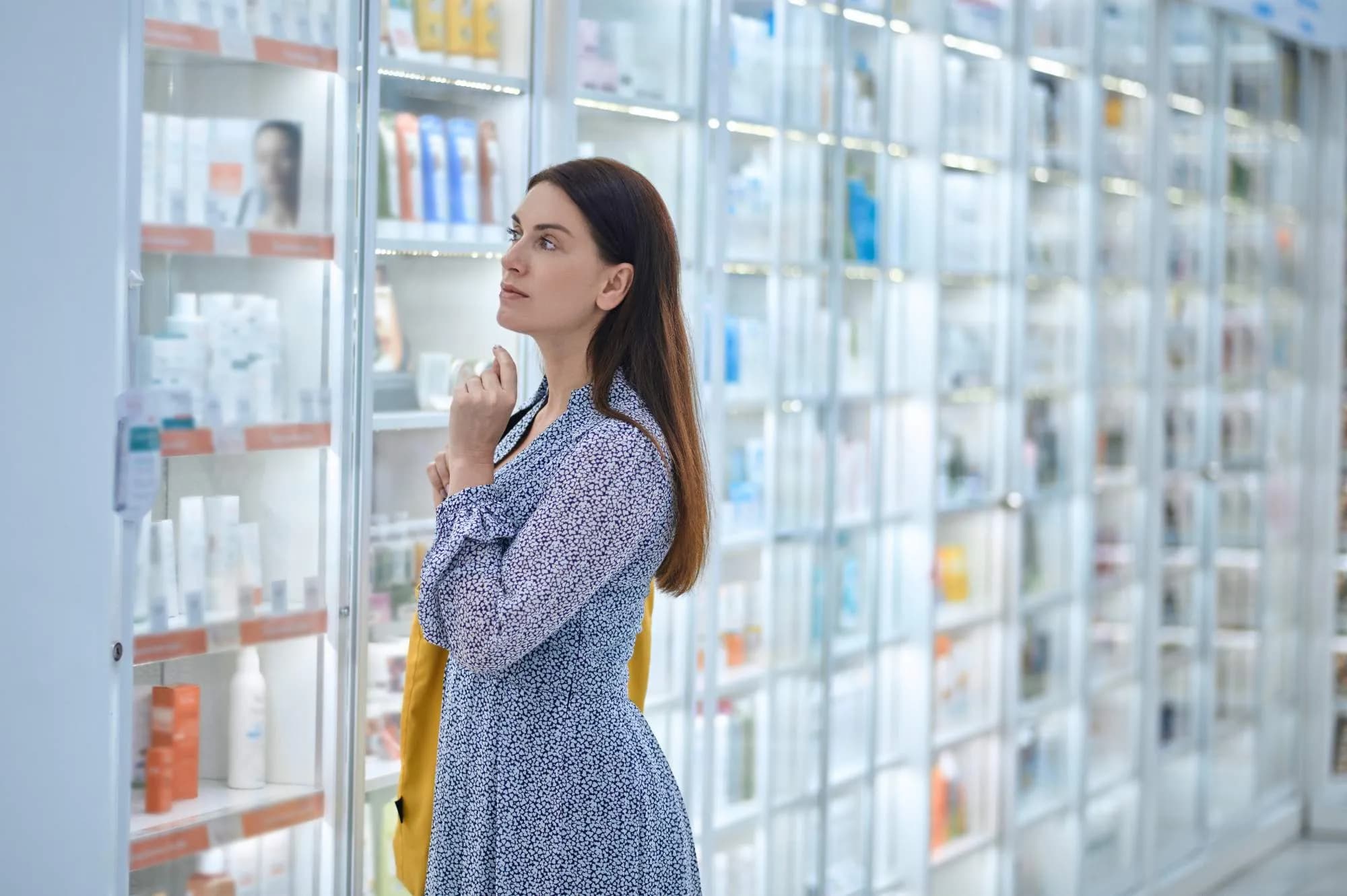 Una dona amb cabell castany llarg, vestida amb un vestit estampat, es troba en una botiga ben il·luminada, mirant atentament els productes exposats en vitrines.
