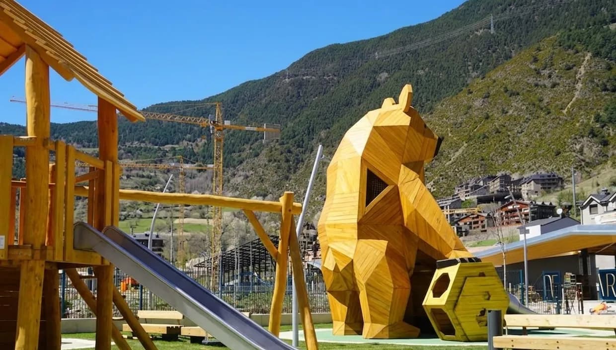 Un parc infantil amb una gran escultura geomètrica d'un os de fusta i una caseta de fusta amb tobogan. Al fons, es veuen muntanyes verdes, edificis i grues de construcció sota un cel blau clar.