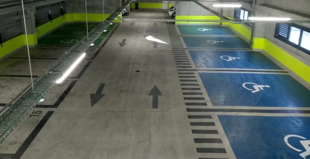 Un estacionamiento subterráneo con plazas de aparcamiento verdes a la izquierda y plazas azules accesibles con símbolos de silla de ruedas a la derecha. Las paredes tienen una franja amarilla brillante, y el suelo de hormigón tiene flechas direccionales y marcas rayadas.