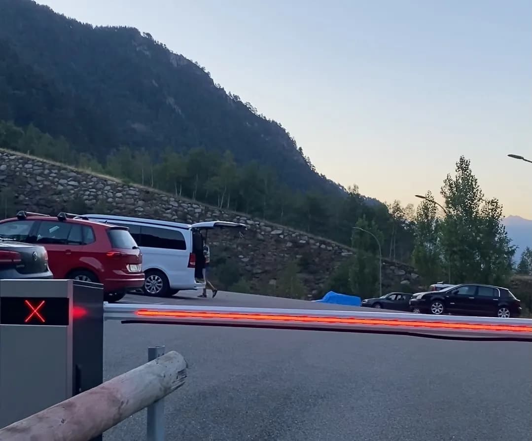 Entrada a un aparcament amb una barrera vermella que mostra una 'X'. Diversos cotxes estan aparcats, incloent un cotxe vermell i una furgoneta blanca amb el maleter obert, amb muntanyes i arbres al fons al capvespre.