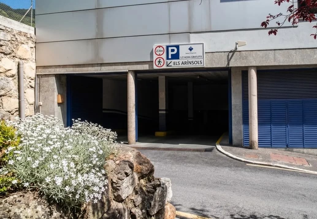 Entrada a un aparcament subterrani amb un rètol que mostra un límit d'altura de 2.30m, '1/2 HORA GRATUITS' i 'ELS ARÍNSOLS'. A l'esquerra hi ha plantes de flors blanques i un mur de pedra.