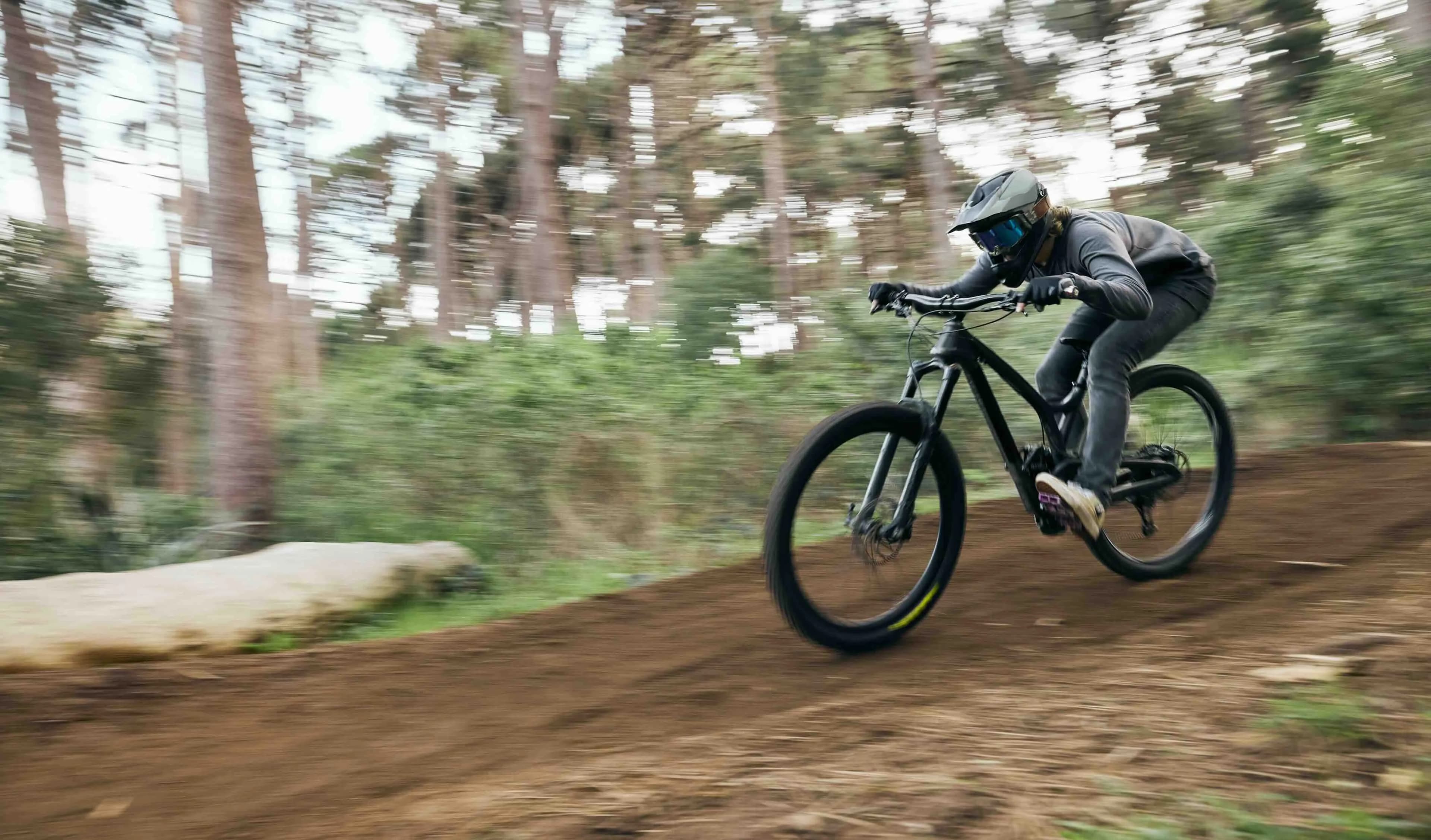 Une personne portant un casque et des lunettes de protection fait du VTT sur une piste de terre à travers une forêt floue, transmettant de la vitesse.